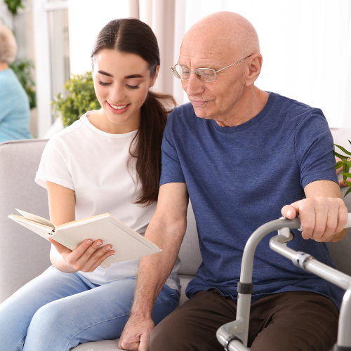 A young woman reads a book to an elderly man sitting beside her.